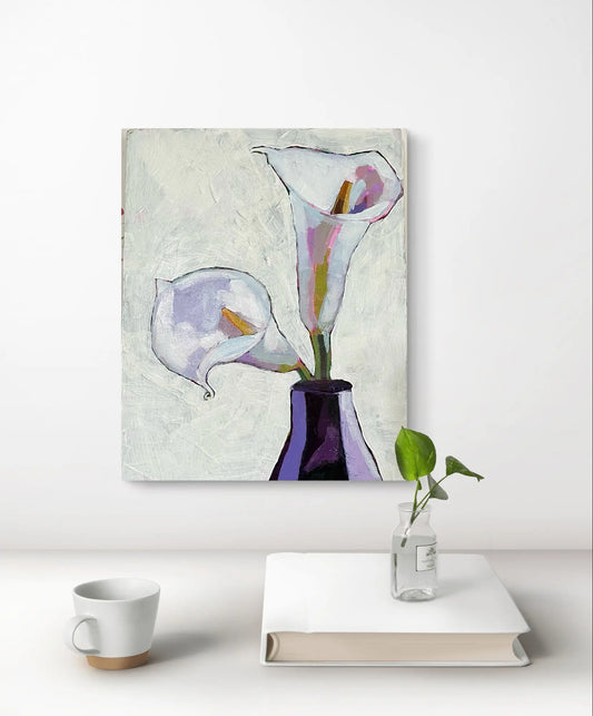 A textured painting of two white and purple calla lilies in a dark vase, displayed on a light wall above a table with a book, small plant, and mug.